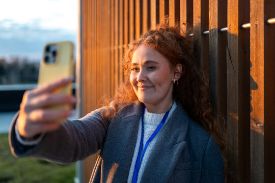 Woman with red curly hair taking selfie on smartphone during golden hour outdoor against wooden wall