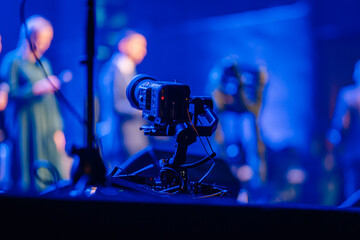 Valmiera, Latvia - December 29, 2025: Professional camera on tripod capturing performers on stage during vibrant blue-lit event with audience in background