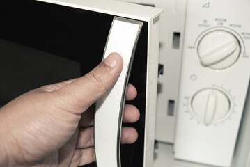Close up of a person's hand opening the door of a white microwave oven. Domestic kitchen appliance and cooking concept.