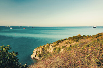 Wide panoramic view of a calm blue ocean with multiple cargo ships on the horizon under clear sky. Rocky cliff and sea scenery in Thailand