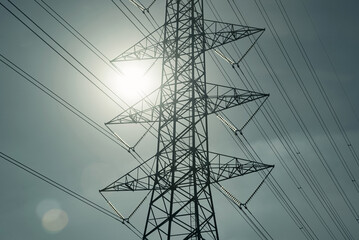 High voltage electric pylon and power lines against a bright sunny sky with lens flare. Electricity infrastructure and energy transmission.