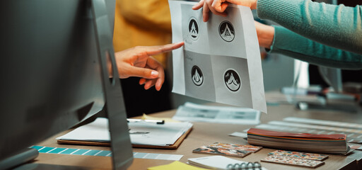 Two creative professionals engage in a collaborative discussion about logo designs. They review printed graphics and share ideas in a bright and modern office environment. SACTR