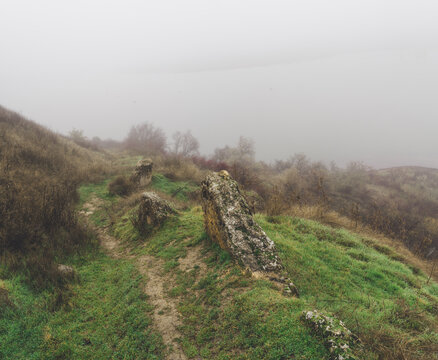 Mysterious landscape with a narrow dirt path on a green grassy hill leading into heavy white fog. Moody autumn morning in nature with ancient stones and dramatic atmosphere.
