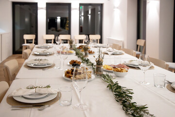 A festive dining table is set with white plates, glasses and rustic greenery, assorted appetizers...