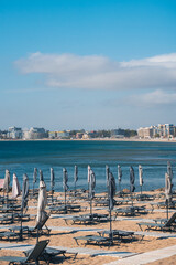 Visitors relax on sunbeds with umbrellas at a beach in Bulgaria. The coastline of sunny beach stretches along the water, while buildings rise in the distance. It is daytime and the sky is clear