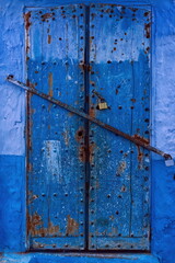 Chipped, dilapidated, studded old wooden door closed with a security bar in the medina, in various paint-faded shades of blue. Chefchaouen-Morocco-034
