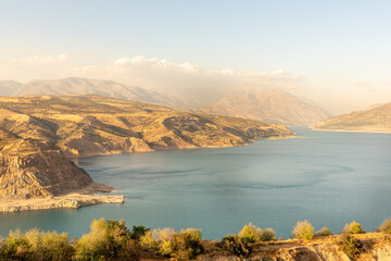 Naklejka premium A landscape shows hills and mountains surrounding a river. The sun shines on the scene during late afternoon. Trees grow near the water, and the sky has few clouds.