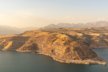 A dry lake sits at the foot of barren hills and slopes. The sun sets in the distance, casting...