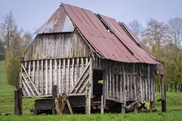 Old abandoned farm barn made of aged wood and corrugated metal, surrounded by countryside greenery. Weathered wooden barn with a rusted metal roof standing in a rural orchard landscape.  © Kaja
