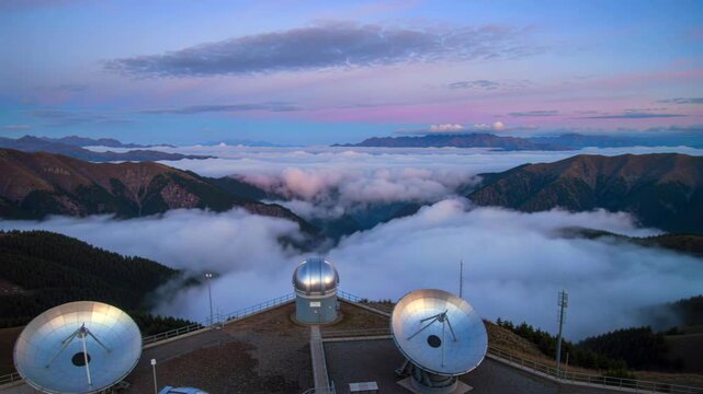 mountain observatory dishes at sunrise, radio telescopes perched on ridge, sea of clouds filling valley, pink and blue sky, mist drifting across peaks, reflective metal dishes, engineers monitoring in