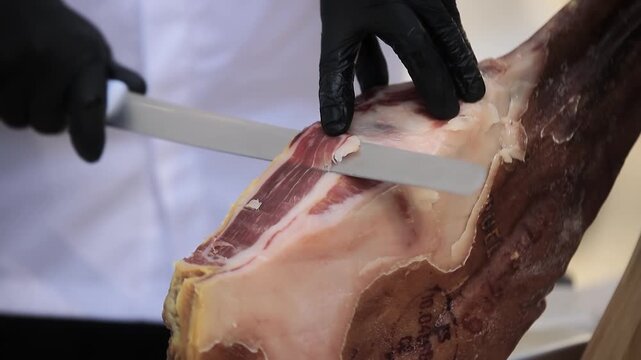 Hands wearing black gloves cut jamon with a long knife. Jamon is cut with a long, sharp knife. A cook wearing black gloves cuts thin slices from a pork leg. Hands wearing black gloves cut thin slices 