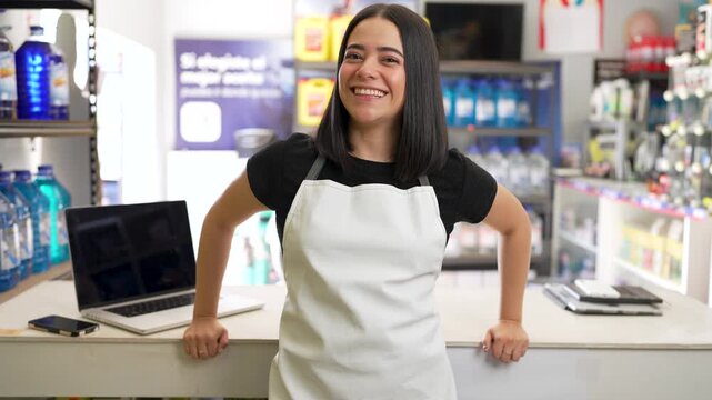 Young hispanic female employee wearing an apron happily smiling while standing behind the counter of a store