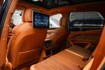 Fototapeta premium Rear cabin of a Bentley Bentayga Speed in an indoor bay shows tan leather, diamond quilted doors, dual rear screens, panoramic roof, and climate console in soft light.