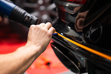 A worker applies and heats a yellow orange vinyl stripe on a glossy black car bumper near the exhaust and tail light, using a squeegee and heat gun in a workshop bay.