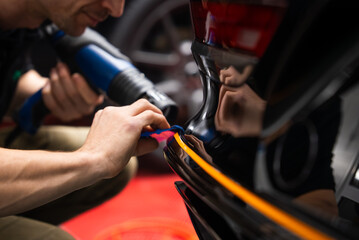 A technician applies an orange vinyl stripe on a glossy black bumper, using a blue squeegee and a heat gun in a workshop, with reflections showing precise work.