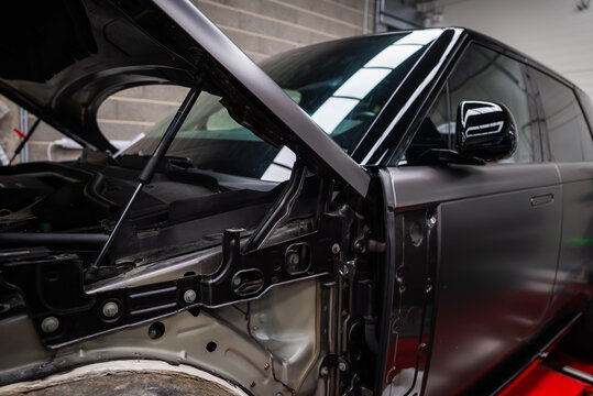 Close view inside an auto workshop shows a matte black SUV mid repair, hood open, fender and wheel arch trim removed, metal and hinge hardware visible under bright lights.