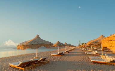 Panoramic shot of a deserted sandy beach with thatched umbrellas and sun loungers during golden hour, with the moon in a blue sky. High resolution