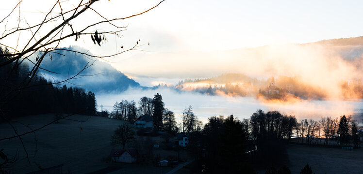 Mari&auml; Himmelfahrt Kirche auf der Insel im See Bled bei Sonnenaufgang mit goldenem Nebel &uuml;ber Kirche und See, ruhige Wasserfl&auml;che und neblige Landschaft im Morgenlicht in Slowenien