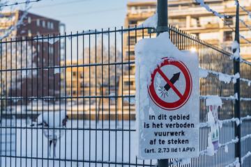 Sign attached to a fence in a Dutch residential area warning that fireworks are prohibited in this zone, covered in winter snow and ice.