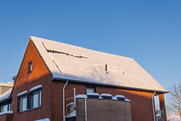 Rooftop of a red brick house in the Netherlands with solar panels partially covered in a thick layer of snow during a sunny winter day