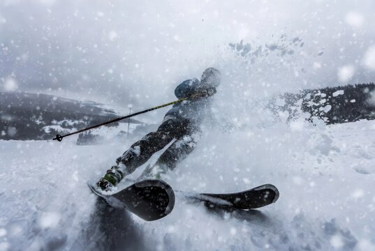 Wide angle close-up of skier falling while carving fast turn with skis crossed and huge spray of fresh powder snow on alpine slope under cloudy winter sky. Concept of risk and intensity in extreme ski