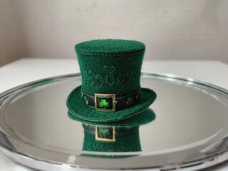 A small green leprechaun hat sits on a reflective surface. The hat features a gold buckle and shamrock designs, perfect for St. Patrick's Day celebrations.
