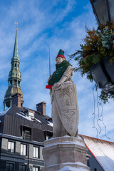 Fototapeta premium A stone knight statue in Riga wears a Christmas tree cloak and elf hat. St. Peter's Church spire rises behind, with modern building and evergreen decor in winter.