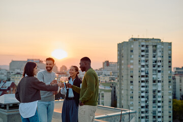 Friends celebrating diversity, laughing, and clinking wine glasses during a rooftop party with a city skyline at sunset