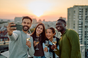 Diverse friends smiling, taking a selfie, and enjoying wine on a city rooftop during a sunset celebration