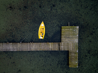 Aerial top-down view of a wooden pier with a small yellow boat moored in clear shallow water. Minimalist coastal scene with strong geometry, calm atmosphere, marine landscape