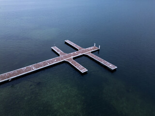 Aerial top-down view of a symmetrical wooden pier over dark water. Minimalist geometric composition with strong lines and contrast, calm atmosphere, abstract coastal structure