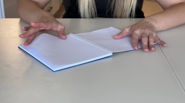 A woman's hand flips through the pages of a blank notepad on a spiral on a brown wooden table, view from the background