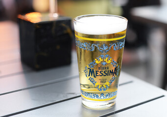 Obraz premium SIENA, ITALY – OCTOBER 7, 2025: Sicilian lager 'Birra Messina' beer in decorated glass. The beer was founded in 1923 and is part of the Heineken group since 1988. Selective focus