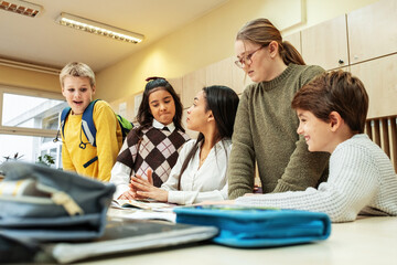 A small group of students is in their elementary classroom while a mixed-race teacher guides them through a math and geometry lesson. She is asking questions and helping them stay focused as they work