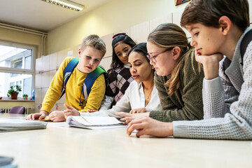 A small group of students is in their elementary classroom while a mixed-race teacher guides them through a math and geometry lesson. She is asking questions and helping them stay focused as they work
