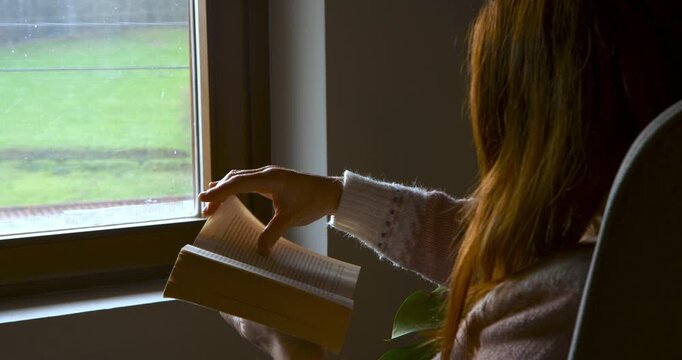A side view of a woman reading a book by a window on a grey day. The movement of the page and the outdoor landscape are visible.