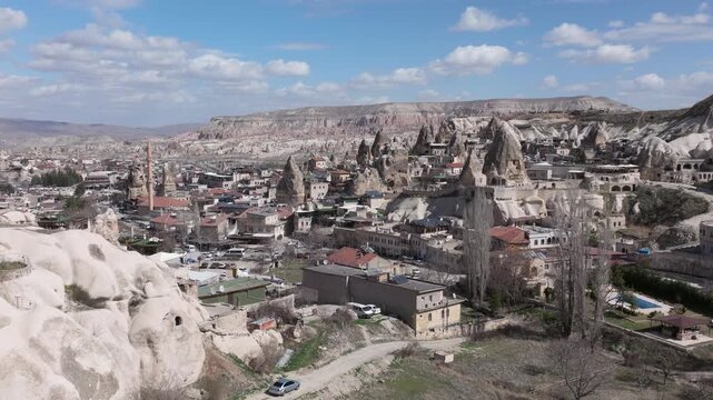 Aerial drone footage of G&ouml;reme town in Cappadocia, Turkey, surrounded by fairy chimneys and volcanic rock formations. The shot reveals traditional stone houses, cave dwellings carved into soft tuff ro