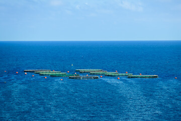 A group of circular offshore fish farm cages surrounded by red buoys floating in deep blue Mediterranean waters under a clear sky.