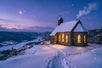 A cozy stone chapel on a snowy hill at twilight. Warm light glows from the windows while snow falls on a peaceful, magical winter Christmas scene.