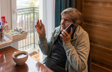 An elderly woman talks on a smartphone and gestures emotionally while sitting at a table near a window. Natural light, authentic lifestyle moment, active communication, modern technology use among sen © Volha