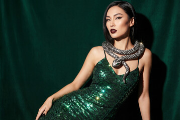 An elegant woman in a sparkling green sequin dress poses with a statement snake necklace against a luxurious, dark emerald velvet background. High fashion portrait.