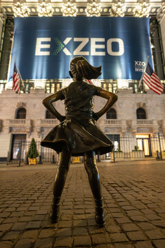 New York, USA - November 24, 2025: The Fearless Girl statue at night, with a brightly lit IPO display of EXZEO in the background, symbolizing courage, ambition, and modern financial markets.