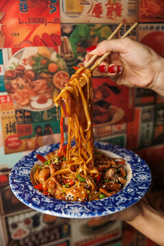 Hand holding chopsticks with noodles and vegetables in a decorative bowl against a colorful food-themed background