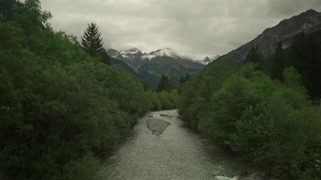 Dramatic view of mountain stream, likely tributary of Inn River, flowing through steep valley near Oberaudorf, Bavaria, Germany. Backdrop is majestic, snow-capped Kaisergebirge (Kaiser Mountains).