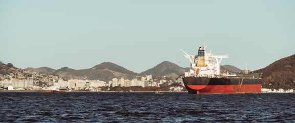 Large cargo tanker anchored in coastal bay with city skyline and green mountains in background under clear sky, calm sea foreground, maritime transport and trade concept, copy space