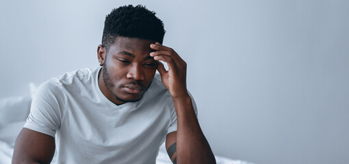 A man is sitting on a bed in a room. He has his hand on his forehead. The light comes from a nearby...