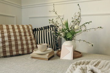 Spring Scandinavian home interior. Still life with coffee cup, books, tulip flowers, blooming branches and greeting card mockup, Minimal nordic living room, sofa with cushions. Calm lifestyle scene