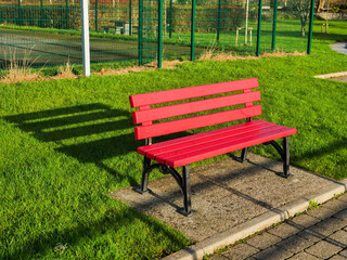 A red bench sits in a grassy area next to a playground fence. The bench is empty and the grass is green