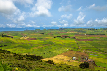 Panoramic view of the colorful agricultural fields from Serra do Cume lookout on Terceira Island, Azores, Portugal. Lush green, yellow, and brown farmlands form a scenic patchwork landscape 