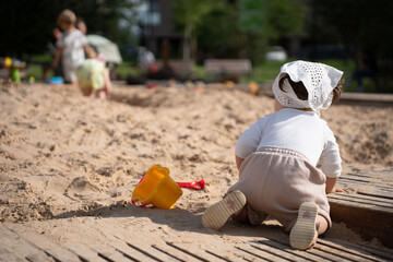 Obraz premium a toddler in a white bonnet crawls in a sunlit sandbox with a yellow bucket and red shovel, surrounded by blurred children playing in a lively park setting, leisure, relax, carefree, day, lively